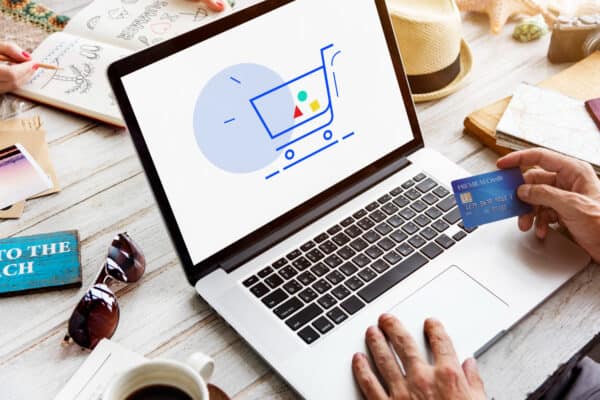 Modernes laptop mit Online-Shopping-Icon und Kreditkarte, geeignet für E-Commerce und digitales Einkaufen, auf stilvollem Holztisch mit Reiseaccessoires und Sonnenbrille.